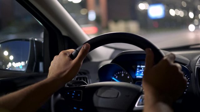 Man driving car in a night city. Male hands holding steering wheel. Oncoming traffic is out of focus with beautiful bokeh. 4K