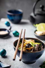 Asian ramen noodles soup with beef, oyster mushrooms and vegetables in bowl on gray background