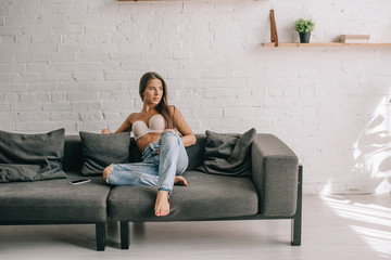 beautiful girl in white bra and jeans sitting on sofa with smartphone