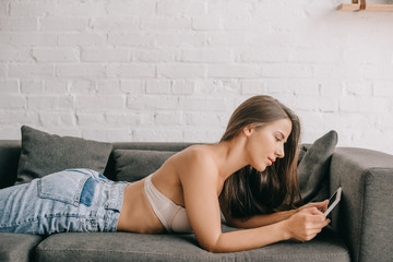 Fototapeta na wymiar beautiful woman in white bra and jeans using digital tablet on sofa in living room
