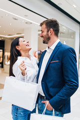 Beautiful couple enjoying in shopping mall or center. They standing in front of huge modern store, smiling and looking at side while holding shopping bags. Shoot from below.