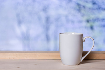 White cup on wooden background at window with blurred snow landscape. Winter comfort concept.