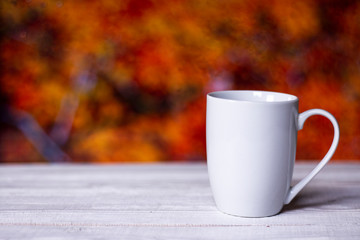 White cup on wooden background at wet window glass with blurred fall colors landscape. Autumn comfort concept.