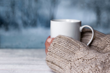 Woman's hands in sweater holding cup with hot drink and blurred winter landscape colors on background. Winter comfort concept.