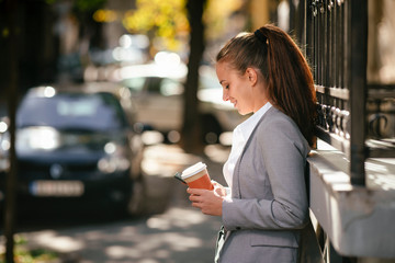 Young woman leaning on wall. Businesswoman drinking coffee and using phone.