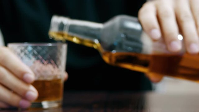 Young drunk man pours and drinks whiskey from a glass and falls asleep on the table, a half-empty bottle of whiskey in the foreground in focus