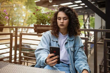 Portrait of a cute brunette curly woman read something on her smart phone while sitting in modern coffee shop. Beautiful young hipster girl using cell telephone while enjoying her free time in cafe.