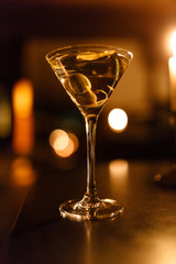 Close up of a cocktail martini with olives on dark night interior with blurry brightness light background. Vertical view.