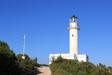 Beacon of Lefkada island south cape stone built lighthouse tower at summer, clear blue sky at background. Pathway and Greek flag at foreground 