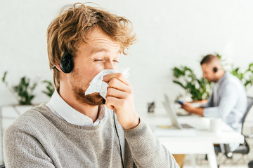 sick bearded broker with closed eyes sneezing in tissue near coworker in office