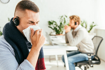 selective focus of sick broker with closed eyes sneezing in tissue near coworker in office