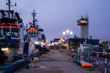 Fototapeta premium EVENING AT THE SEAPORT - Sleeping tugs at the port quay against the backdrop of a 24-hour watchful sea control tower