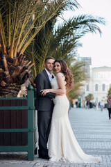 Beautiful couple posing on streets near palm trees. Young sexy couple hugs near palm trees, enjoy their romantic vacation together. Couple wearing suit and white luxury dress, enjoy their honeymoon