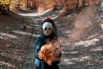 Photo of  walking girl with creepy skull mask in the autumn woods, holding a carved smoky Halloween pumpkin.  On background path in autumn forest with fall leaves. Concept about Halloween.