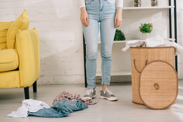 cropped view of housewife in denim jeans standing near laundry basket and clothes on floor