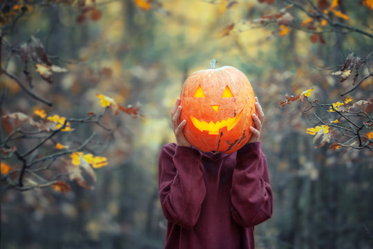 Boy holding carved pumpkin for halloween in front of his head in dark autumn forest.