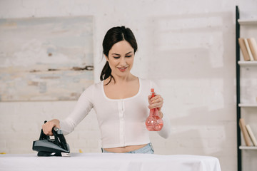 smiling housewife holding spray bottle and holding iron while standing near ironing board