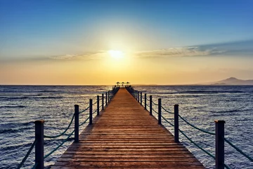 Foto auf Acrylglas Heller und farbenfroher Sonnenaufgang über dem Meer und dem Pier. Perspektivischer Blick auf einen Holzsteg am Meer bei Sonnenaufgang mit felsigen Inseln in der Ferne © RealVector