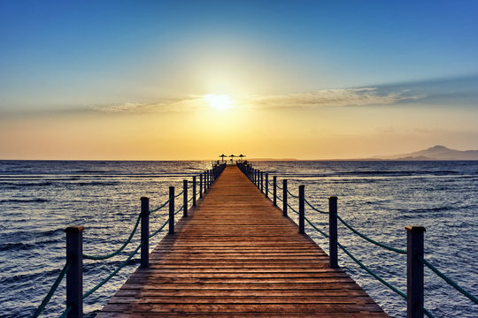 Bright and colorful sunrise over the sea and pier. Perspective view of a wooden pier on the sea at sunrise with rocky islands in the distance