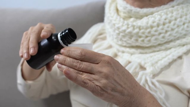 Old woman in scarf taking cough syrup and smiling at camera, effective medicine