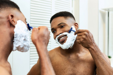 Cheerful black guy shaving in bathroom, looking at mirror