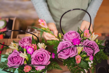 Florist girl with her hands makes bouquet flowers of purple roses, fern in gift box. Top view on workplace