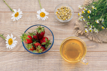 Glass cup of green tea, stawberries with white chamomile flowers on wooden background.