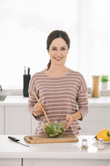 happy young woman looking at camera while mixing fresh vegetable salad