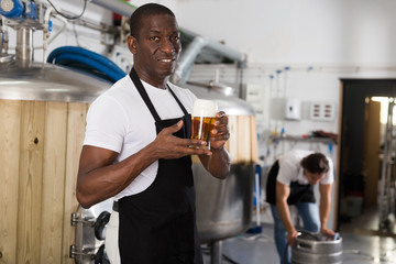 African american man brewer in apron standing with glass of beer