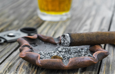 On an old wooden table lies a smoldering cigar on an ashtray. In the background a defocused...