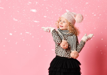 Little baby girl in warm hat and gloves standing under snow, holding something on her open palms on pink with copy space