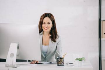 attractive, smiling secretary looking at camera while sitting at workplace
