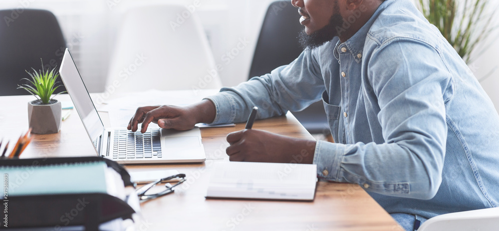 Poster Black trainee taking notes while using laptop in office