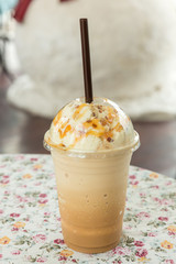 Ice coffee with cream on table. Caramel frappe coffee in the coffee shop. Iced blended frappucino. Frappuccino in plastic takeaway or to go cup on wooden table at cafe