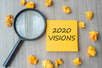 2020 VISIONS words on yellow note with crumbled paper and magnifying glass on wooden table...