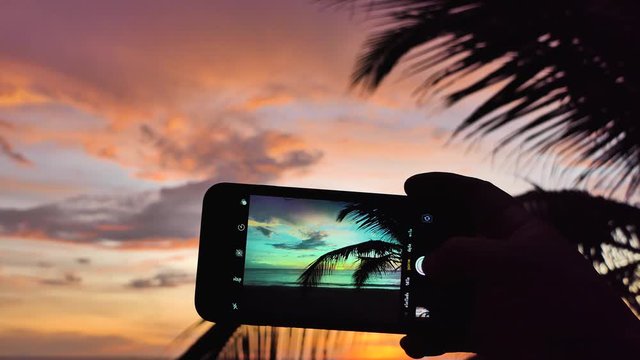 The hand of a man holding a smartphone and taking a picture of the sea at sunset
