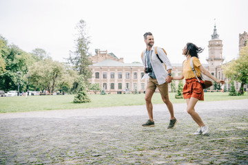 Obraz premium cheerful bearded man and woman holding hands while running near building