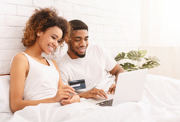 Happy african couple shopping online in bed, using laptop and credit card