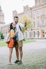 happy bearded man and attractive young woman taking selfie near building