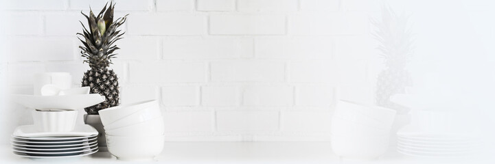 Piece of kitchen interior with white porcelain dishes, kitchen table and pineapple fruit. Copy space. Wide panoramic white foggy food background.