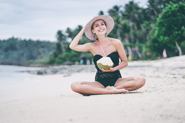 Vacation on the seashore.Young woman in hat and swimsuit holding green cococnut relaxing on the white sand beach.