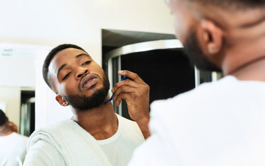 Young black man shaving beard, looking at bathroom mirror
