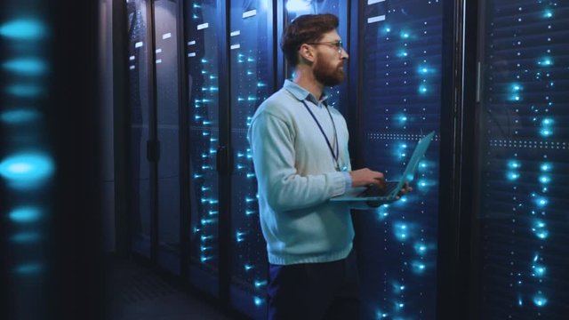 Smart IT specialist with notebook controlling rack server cabinet in big database center for diagnostics working at render farm. Digitalization of information.