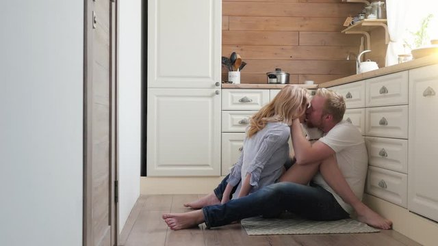 pregnant wife sitting on the floor with her husband. a man and a pregnant woman are sitting at the back of their kitchen and kissing. Scandinavian style.