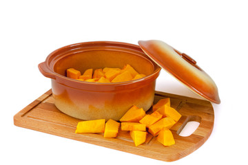 Fototapeta premium A small ceramic pan with an open lid filled with sliced pumpkin on a wooden cutting board on which pieces of pumpkin also lie. Isolated On white background.