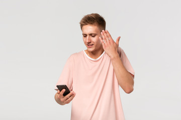 Confused blond teenager in a pink tee looking at the screen of smartphone isolated over light grey background.