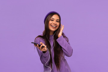 Pretty african-american girl in a hat and sweater dancing listening to music on a mobile phone with wired headphones isolated over purple background.
