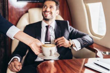 cropped view of flight attendant giving cup of coffee to handsome businessman in suit in private plane