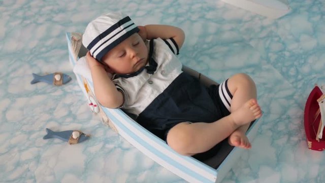 Cute toddler baby boy sleeping in wooden boat with fishes, starfish and binoculars, dressed as sailman, indoors shot