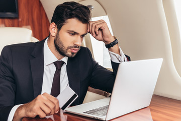 handsome businessman in suit holding credit card and using laptop in private plane
